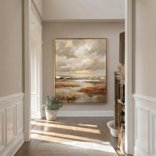 A framed landscape painting hangs on a beige wall in a bright hallway with white wainscoting, sunlight streaming in from an open doorway, and a small potted plant nearby.